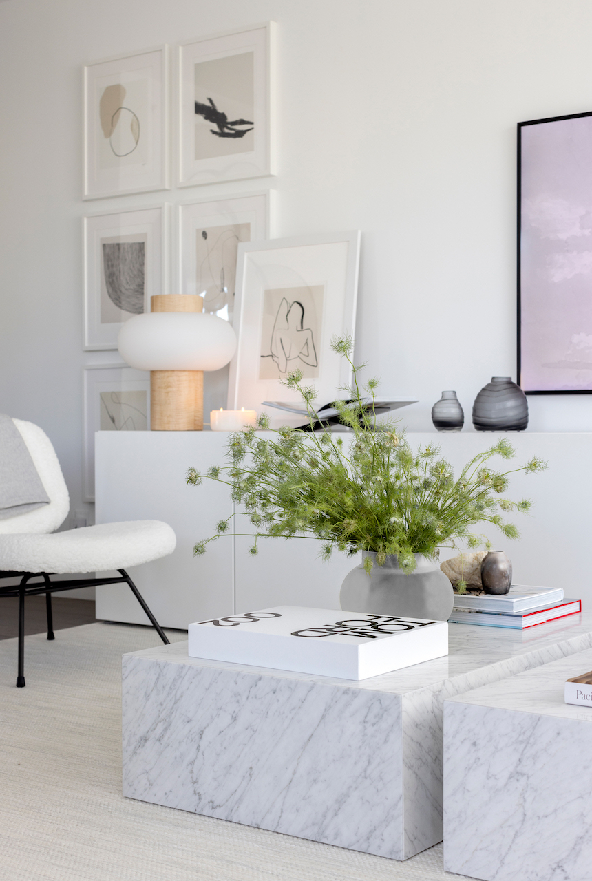 white interior of living room with marble coffee table and oversized green flower arrangement gallery wall in the background with white fluffy chair and black legs