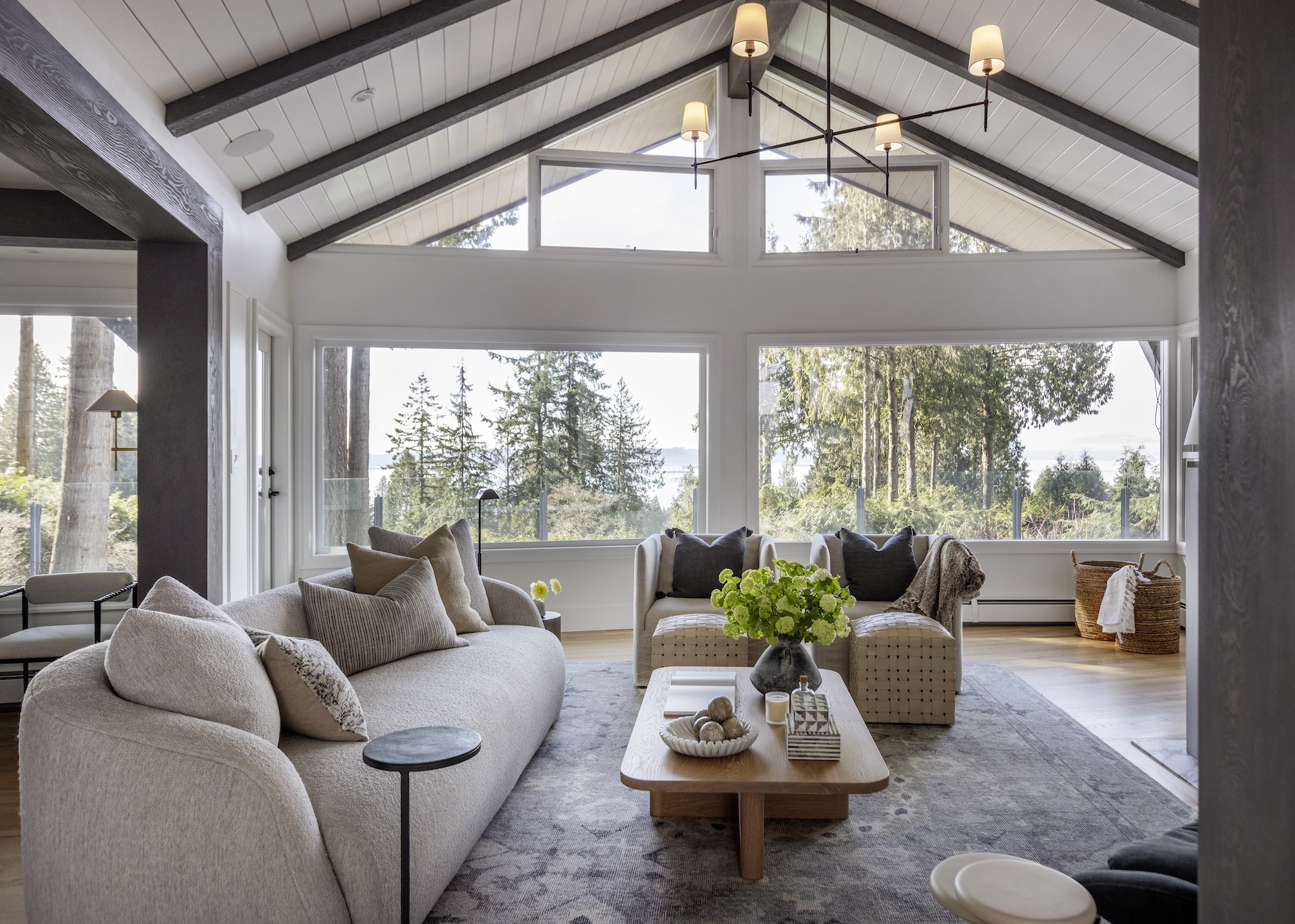 luxury interior design megan baker interiors west coast reno layered living room with peaked ceiling grey beams white shiplap betwee large windows with a view of the forest and ocean behind neutral grey blue charcoal and beige boucle sofa with soft pillows white oak coffee table decorated with natural bone marble and clay accents bright green flowers in the middle floral pattern rug with subtle colour palette
