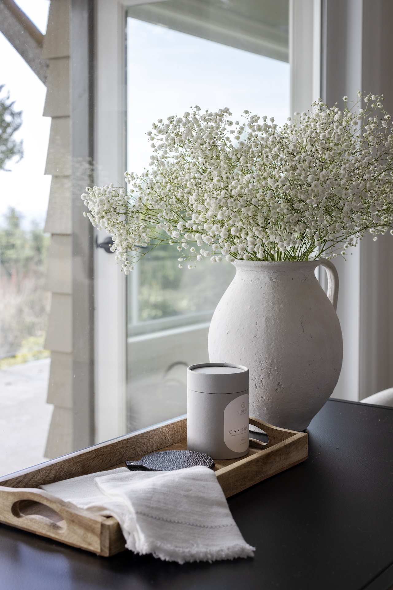 megan baker interiors luxury interior design west coast reno antique olive jug vase with white babys breath overflowing wood tray with linen napkins and leather coaster on it candle in a soft grey box linnea brand