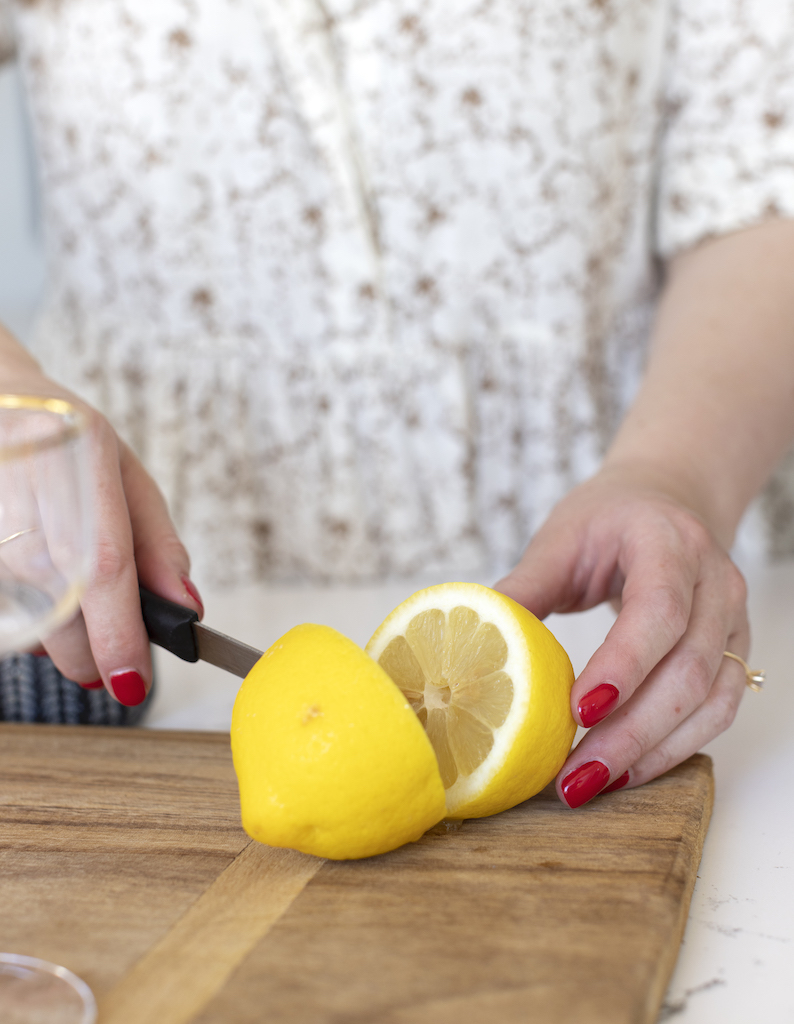 lemon being sliced for sidecar cocktail recipe, bright, close up image of lemon featuring hands and a cutting board