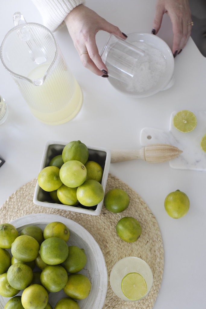 ingredients for a margarita birds eye view. bright green limes, woven placemat, wood lime juicer, hands holding glass